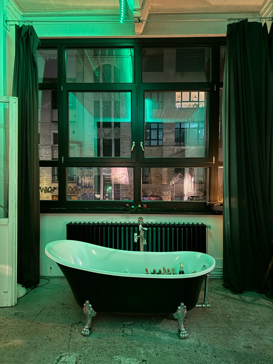 Freistehende Badewanne vor dem großen Loftfenster in grünem Licht