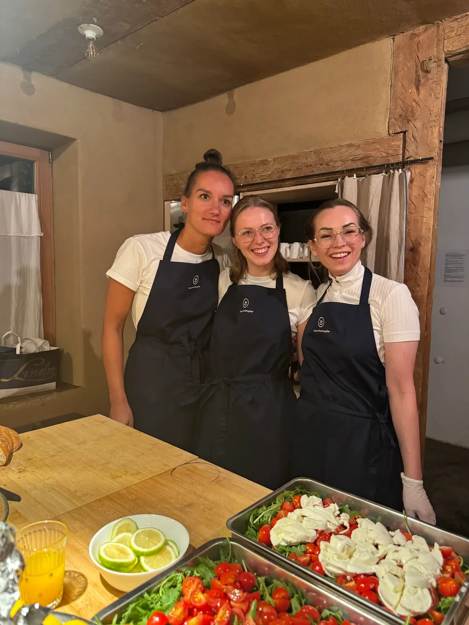 Drei Kolleginnen in Schürzen lächeln hinter Blechen mit Tomaten und Mozzarella