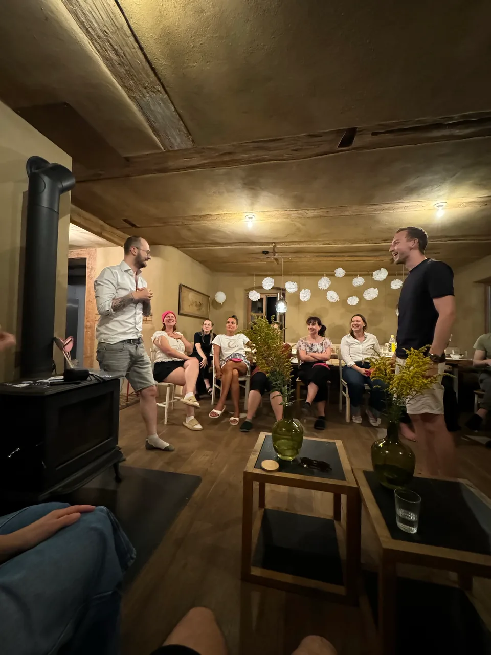 Runde im Wohnzimmer lacht, zwei Kollegen stehen in der Mitte und sprechen